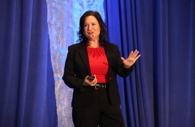 Woman in black blazer and red blouse speaking on stage with blue curtains in background